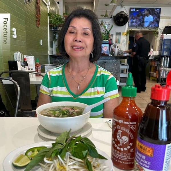 A woman smiles at a table with a bowl of pho, fresh herbs, and sauces at Pho Viet Cali.