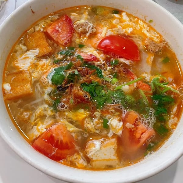 Close-up of a vibrant Vietnamese soup with tomatoes, tofu, shrimp, and herbs in a white bowl.