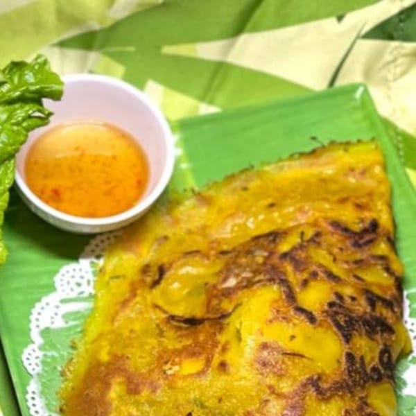 Crispy Vietnamese bánh xèo pancake with a side of dipping sauce and lettuce on a green plate.