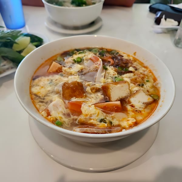 A bowl of Vietnamese soup with tofu, egg, and vegetables on a white table.