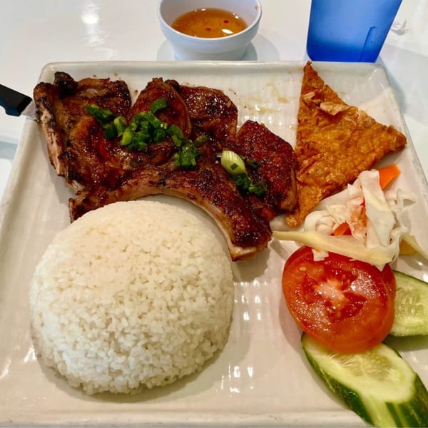 Grilled pork chop with rice, fried egg roll skin, and fresh vegetables on a white plate.
