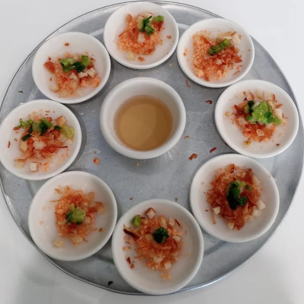 A round metal tray holds eight small white bowls of Vietnamese Banh Beo with a central dipping sauce.