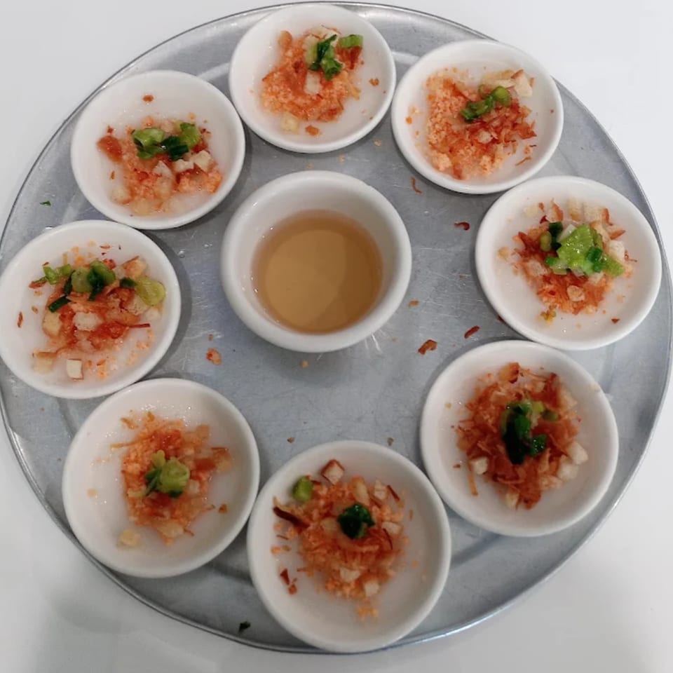 A round metal tray holds eight small white bowls of Vietnamese Banh Beo with a central dipping sauce.